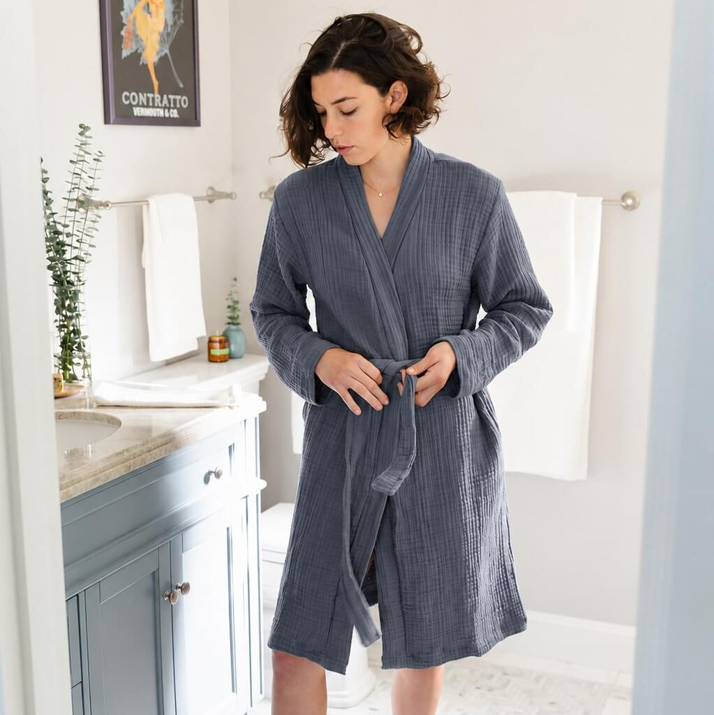 A woman wearing a slate blue-gray wrap robe and mauve socks standing in a bright bathroom doorway.
