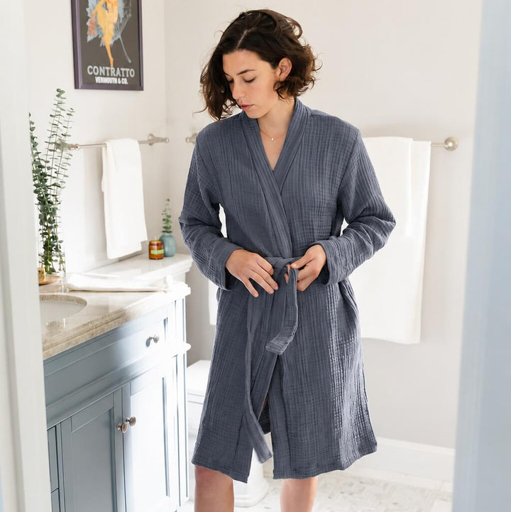 A woman wearing a slate blue-gray wrap robe and mauve socks standing in a bright bathroom doorway.