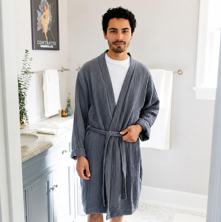A man wearing a slate colored bathrobe over a white t-shirt and white socks, standing in a bright bathroom.