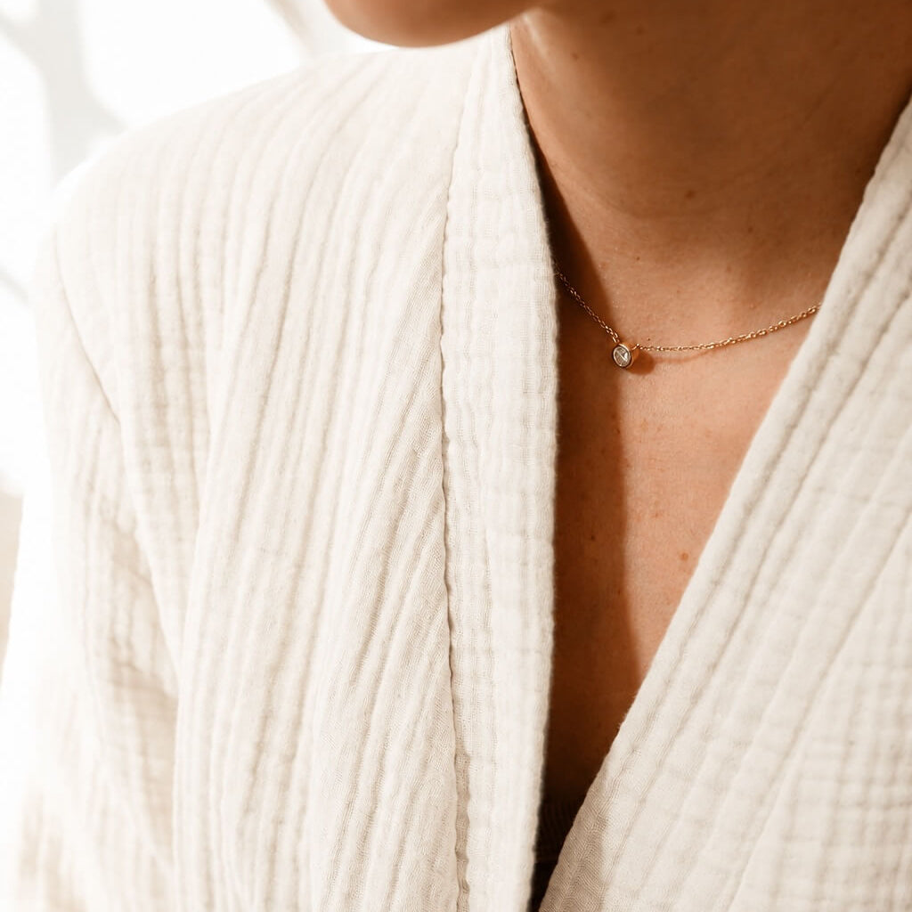 Close-up of a person wearing a cream-colored textured cotton robe and a delicate gold chain necklace with a small bead pendant.